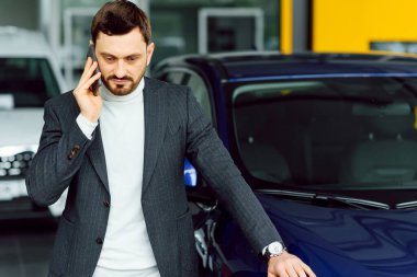 Araba bir adamın karakterini gösteriyor. Kendine güveni tam, şık takım elbiseli ve elinde telefonla yeni mavi arabanın yanında duran zarif iş adamı başka tarafa bakıyor.
