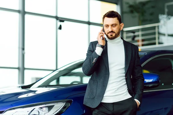 The car shows the character of a man. Confident success elegant business man in full suit and with phone in hand standing near new blue car looks away