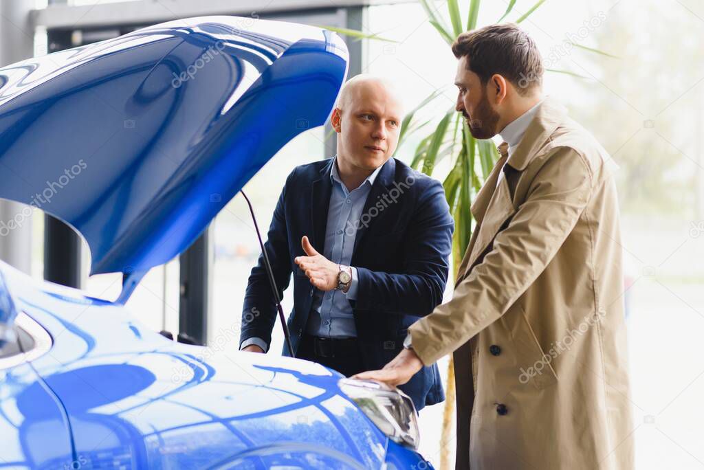 Car dealer and his ale client looking under the hood of a new car on sale at the dealership. Young man buying auto at car salon