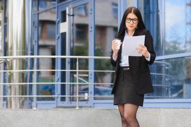 Ofis binasının yakınındaki kahve dükkanında güzel bir kadın işe gidiyor. Başarılı bir iş kadınının portresi. Şehir caddesinde işe giderken elinde sıcak bir bardak içki tutuyor. Yüksek Çözünürlük.