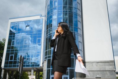 Gözlüklü, koyu renk ceketli ve mavi gömlekli genç bir ofis çalışanı ofis binasının yanındaki caddede kalıyor ve belgelerini tutuyor..