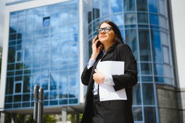 Ofis yakınında telefonu olan bir iş kadını. Moda içinde gülümseyen güzel bir kadının portresi. Ofis kıyafetleri dışarıda dururken telefonda konuşuyor. Telefon iletişimi. Yüksek Kalite Resim.