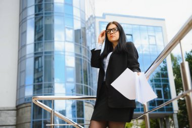 Gözlüklü, koyu renk ceketli ve mavi gömlekli genç bir ofis çalışanı ofis binasının yanındaki caddede kalıyor ve belgelerini tutuyor..