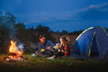 Dağlarda kamp gecesi. Yanan kamp ateşiyle yanan çadırın önünde oturan turist çift. Turizm ve açık hava faaliyeti kavramı.