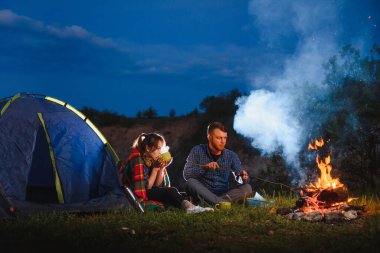 Genç çift kahve içiyor ve alacakaranlıktaki orman tepesinde kamp ateşinde ısınıyor. Aktif dinlenme ve çadır ile seyahat kavramı.