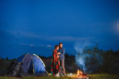 Dağlarda gece kampı. Kamp ateşi ve parlayan turist çadırının yanında birlikte oturan mutlu çiftler. Arka planda büyük kaya, orman ve gece gökyüzü.
