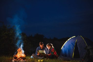 Dağlarda kamp gecesi. Yanan kamp ateşiyle yanan çadırın önünde oturan turist çift. Turizm ve açık hava faaliyeti kavramı.