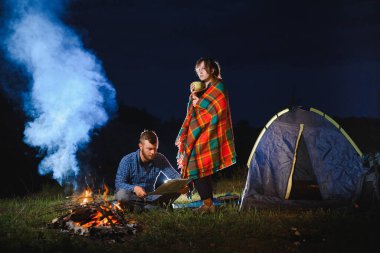 Birbirini seven çift yürüyüşçüler, gece kamp ateşinin yanında, gökyüzünün altında ağaçların ve çadırların yanında. Dağlardaki ormanın yakınında romantik bir kamp.