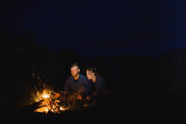 Dağlarda gece kampı. Kamp ateşi ve parlayan turist çadırının yanında birlikte oturan mutlu çiftler. Arka planda büyük kaya, orman ve gece gökyüzü.