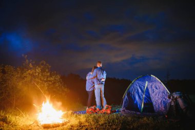 Genç çift ve kadın trevelers parlayan turist çadırının yanında oturan, kamp ateşi yakan, dağın tepesinde, gece gökyüzünün güzel manzarasının tadını çıkaran.