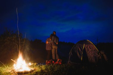 Birbirini seven çift yürüyüşçüler, gece kamp ateşinin yanında, gökyüzünün altında ağaçların ve çadırların yanında. Dağlardaki ormanın yakınında romantik bir kamp.