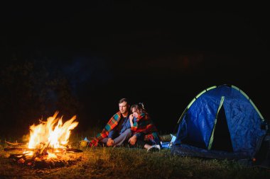 Genç çift ve kadın trevelers parlayan turist çadırının yanında oturan, kamp ateşi yakan, dağın tepesinde, gece gökyüzünün güzel manzarasının tadını çıkaran.