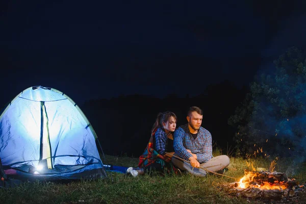 Night camping in the mountains. Happy couple travellers sitting together beside campfire and glowing tourist tent. On background big boulder, forest and night sky.