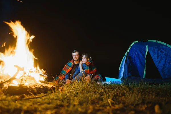 Night camping in the mountains. Happy couple travellers sitting together beside campfire and glowing tourist tent. On background big boulder, forest and night sky.