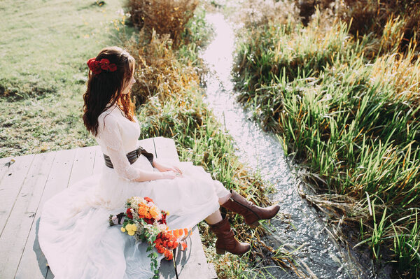Beautiful bride with a bouquet and in boots sits on a bridge against the background of the lake