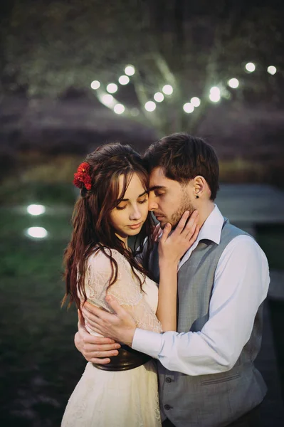 La novia y el novio posando en la noche sobre el fondo de luces en la naturaleza. abrazó a la novia —  Fotos de Stock
