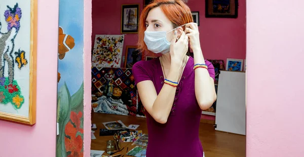 A young slender girl, an artist, in a medical mask stands in front of a mirror. Reflection in the mirror. Coronavirus quarantine self-isolation period.