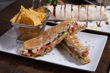 İtalyan ciabatta sandviçi, tavuklu, domatesli ve beyaz tabakta nachos.
