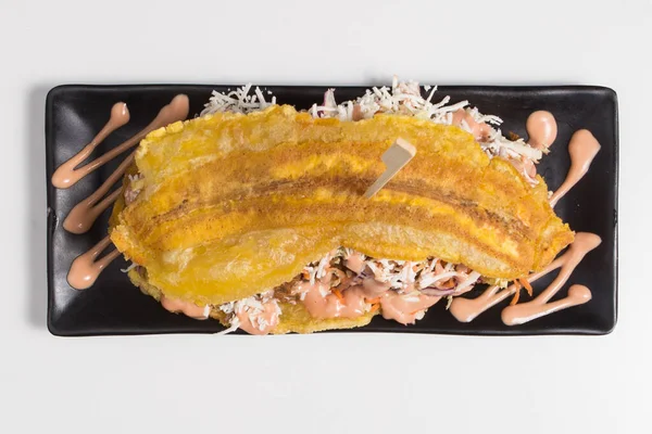 COLOMBIAN CARIBBEAN CENTRAL AMERICAN FOOD. Patacon or toston, fried and flattened whole green plantain banana on white plate with tomato sauce and chicharron white background, top view