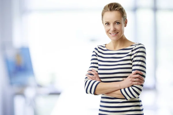 Smiling middle aged sales manager woman standing with arms crossed in office.
