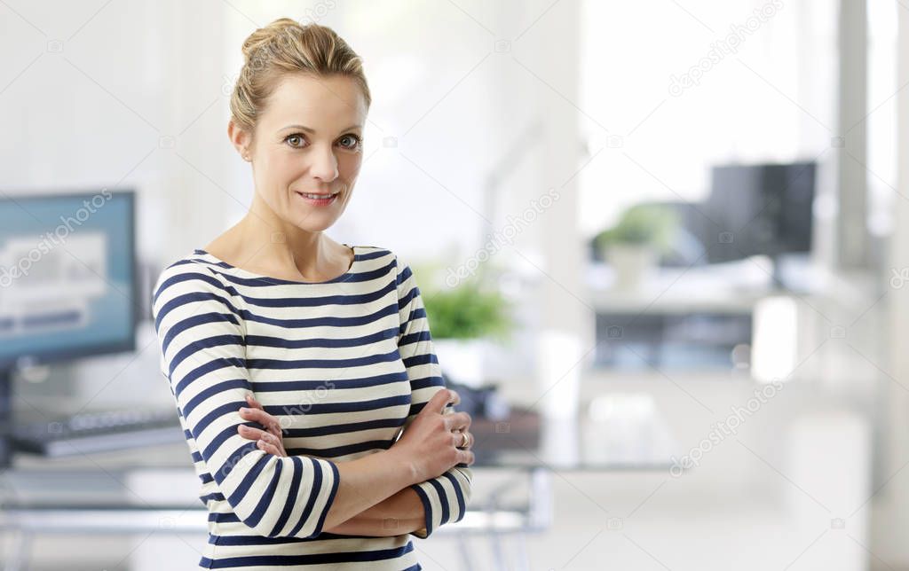 Smiling middle aged sales manager woman standing with arms crossed in office. 