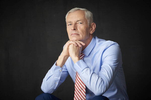 Elderly man looking thoughtfully while sitting before dark grey wall with hand on his chin.