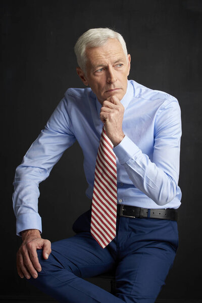 Confident senior man wearing shirt and tie and looking thoughtful while sitting before dark grey wall.
