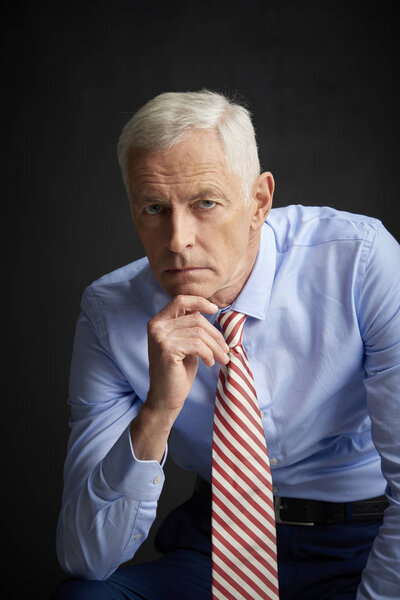 Confident senior man wearing shirt and tie and looking thoughtful while sitting before dark grey wall.