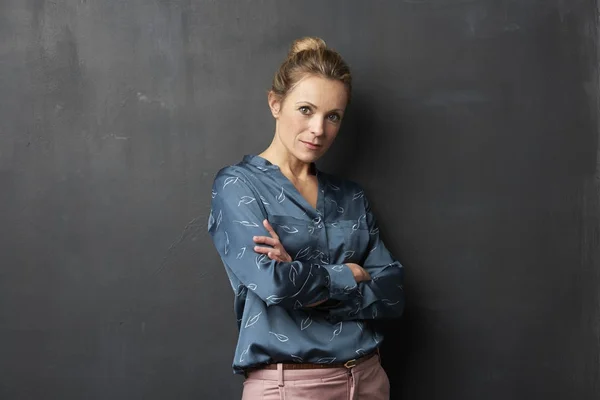 Attractive middle aged woman standing with arms crossed at dark grey wall.