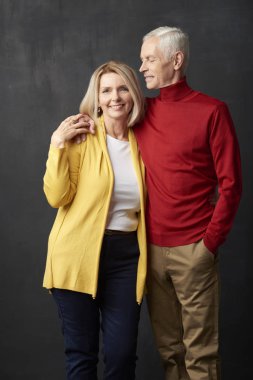 Koyu arka plan, mutlu üst düzey çift ayakta. Kameraya bakarak ve yaşlı adam onu kucaklayan gülümseyen güzel Olgun kadın.