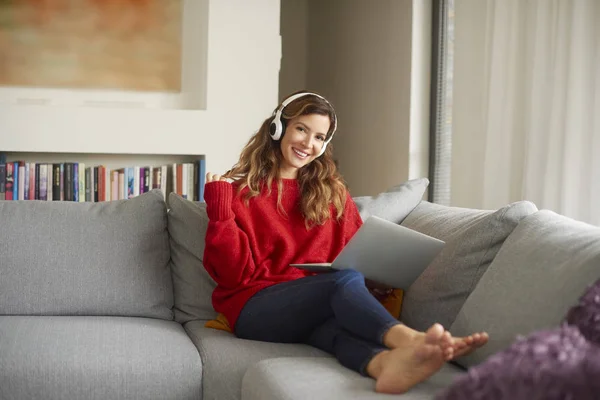 Full length shot of attractive woman with headphone and laptop sitting on sofa and listing to music.