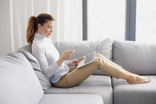 Full length shot of attracitve woman using mobile phone and laptop while working from home.