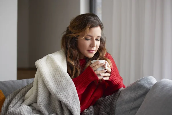 Mature woman relaxing at home with cup of coffee while sitting on sofa and she covers herself with a blanket.