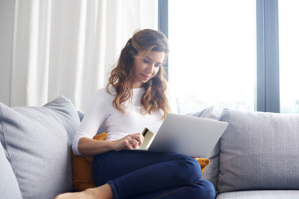 Beautiful middle aged woman using credit card and laptop while banking online from her comfy sofa.