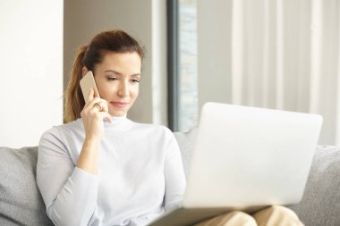 Dizüstü bilgisayar ve çalışma ofisten kanepede otururken arama yapma ve cep telefonunu kullanarak çekici orta yaşlı kadın.