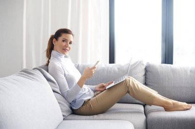 Tam uzunlukta atış bir kanepe oturma ve cep telefonu ve laptop süre çalışma evden kullanarak çekici Olgun kadın. 