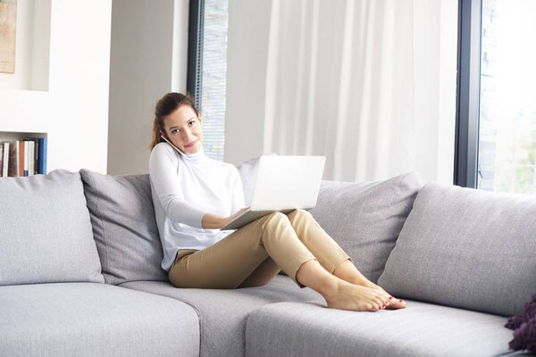 Full length shot of an attractive mature woman sitting a sofa and using mobile phone and laptop while working from home. 