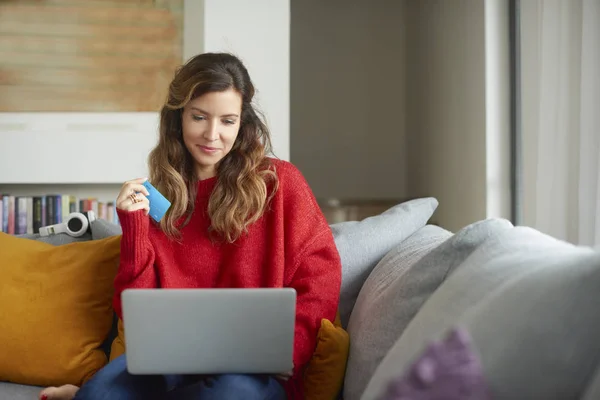 有魅力的成熟女人坐在沙发上在家 拿着她的银行卡和使用笔记本电脑 而支付账单在线 — 图库照片