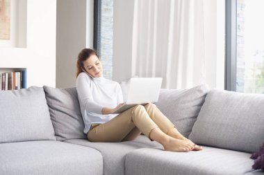 Tam uzunlukta olan dizüstü bilgisayar ve çalışma ofisten kanepede otururken arama yapma ve cep telefonunu kullanarak çekici orta yaşlı kadın.