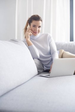 Cep telefonu kullaranarak ve laptop ve evden çalışan ile kanepede otururken arama yapma çekici kadın gülümsüyor. Ev ofis. 