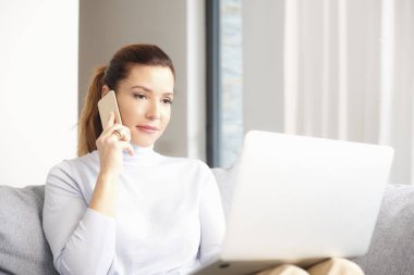 Cep telefonu kullaranarak ve laptop ve evden çalışan ile kanepede otururken arama yapma çekici kadın gülümsüyor. Ev ofis. 