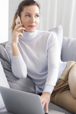 Cep telefonu kullaranarak ve laptop ve evden çalışan ile kanepede otururken arama yapma çekici kadın gülümsüyor. Ev ofis. 