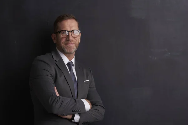 Portrait shot of a smiling middle aged man wearing suit and standing with arms crossed at dark background.