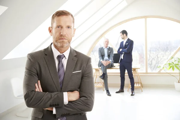 Businessman with folded arms standing at the office. Professional man wearing suit and looking at camera while his unrecognizable collegue consulting at the background.
