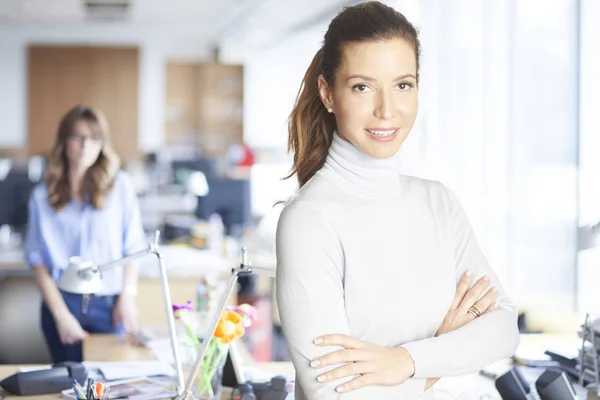 Successful mature sales woman standing with arms crossed at the office and looking at camera while her colleague standing in the background.