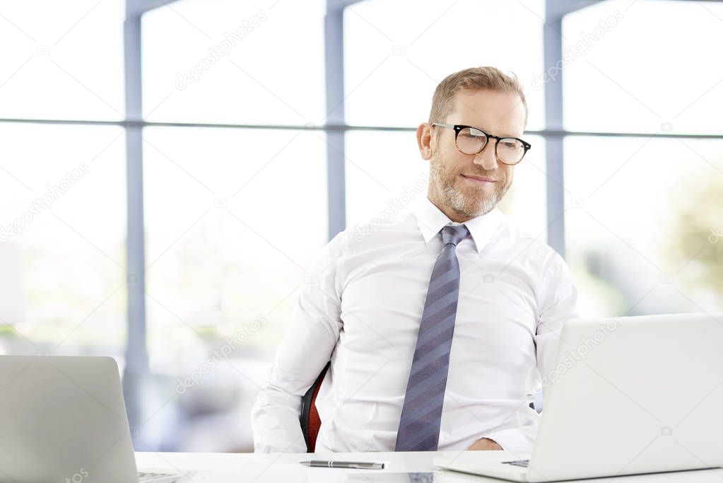 Portrait of middle aged senior agent wearing shirt and tie while sitting at desk and working on laptop at the office. 