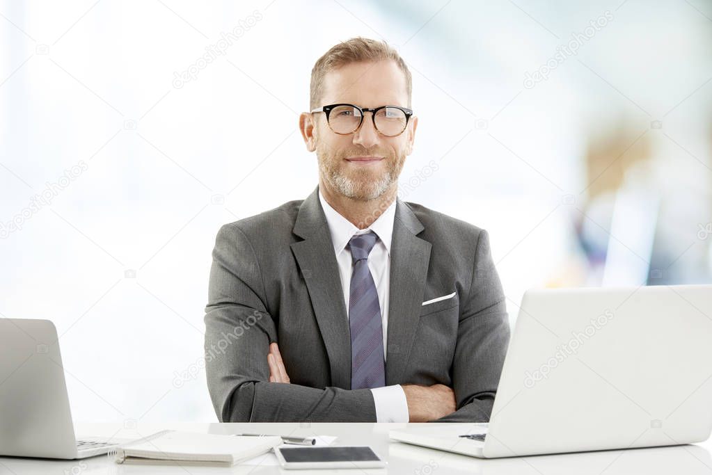 Executive businessman with arms crossed sitting at the office desk and looking at camera.