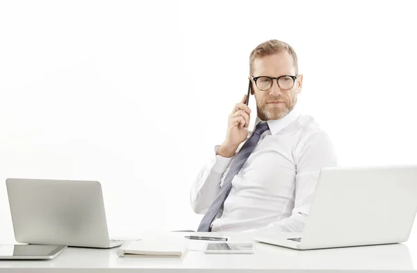 中年男性のビジネス ブローカーの携帯電話を使用して ラップトップの前の机に座っている間の呼び出しを行います 白い背景に分離 — ストック写真