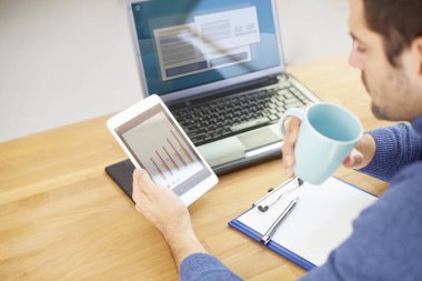 Rear view shot of young businessman holding digital tablet in his hand and analyzing financial data while sitting at desk in front of laptop and drinking coffee. Focus on foreground.  clipart