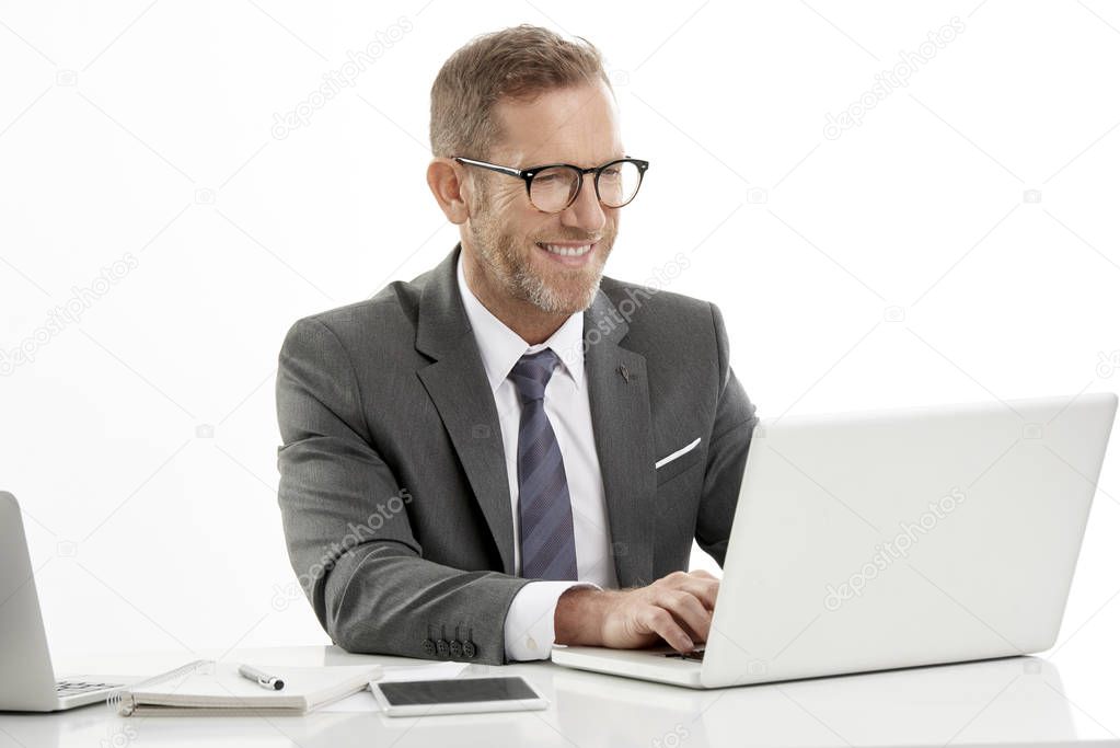 Studio shot of middle aged businessman wearing suit and working on laptop while sitting at desk. Isolated on white background. 
